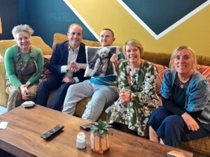 PSS Shared Lives carer, Sally Smith, MP Aaron Bell, supported person Paul with PSS UK CEO, Lesley Dixon and Registered Manager Lisa Hill. They sit on an extra large couch together and Paul is holding up a pillow with a picture of their dog on it.